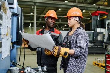 Two manufacturing workers wearing protective gear review a large work plan together on the production floor