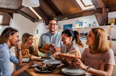 Coworkers sitting together and sharing pizza during a casual team meal in a bright workplace setting.